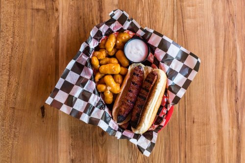 A basket with two grilled hot dogs, cheese puffs, and a small cup of dipping sauce on a checkered paper.