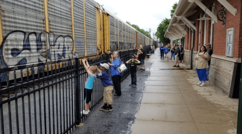 A group of people watch a train pass by while standing along a fence on a rainy day.