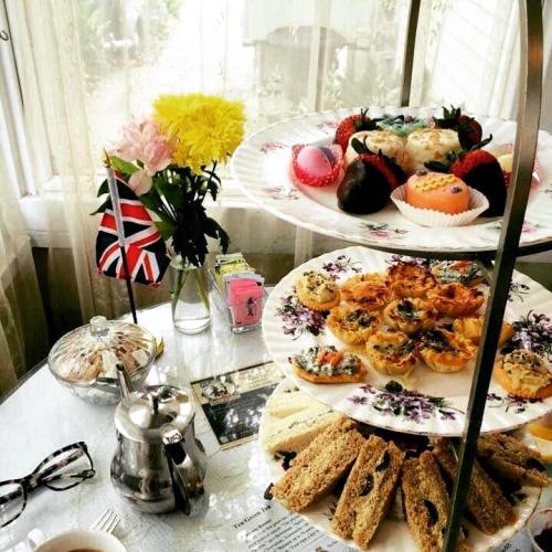 A tiered stand of assorted pastries and snacks, with flowers and a teapot on a table in a bright, cozy setting.