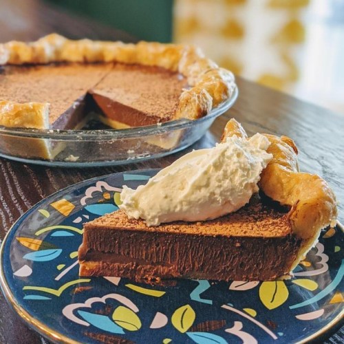 A slice of chocolate pie topped with whipped cream on a decorative plate, with the whole pie in the background.