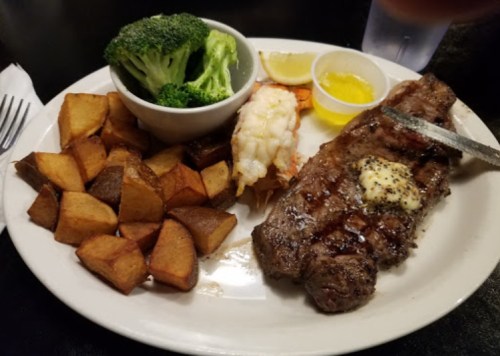 A plate featuring grilled steak with butter, shrimp, broccoli, and seasoned potato wedges, served with lemon and dipping sauce.