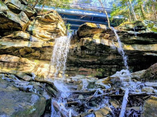 A scenic view of a waterfall cascading over rocky cliffs, with icicles and a bridge above surrounded by trees.