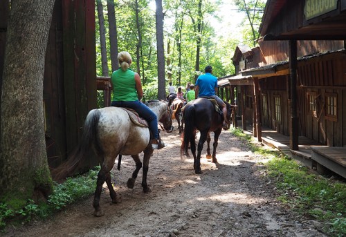 Two riders on horseback walk down a dirt path lined with trees and rustic buildings in a wooded area.