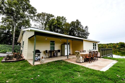 A cozy lakeside cabin with a patio, outdoor dining set, and grill, surrounded by trees and greenery.