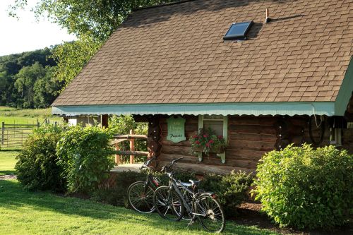 A cozy log cabin with a green awning, surrounded by greenery and two bicycles parked in front.