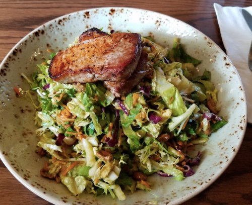 A bowl of mixed salad topped with a seared piece of meat, featuring greens, cabbage, and crunchy toppings.