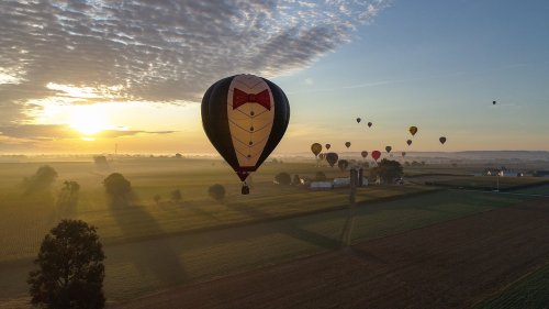 A colorful hot air balloon with a bow tie design floats over a rural landscape at sunrise, surrounded by other balloons.