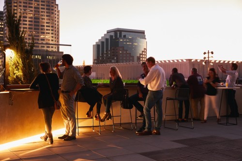 A rooftop bar scene at sunset with people socializing and enjoying drinks against a city skyline backdrop.