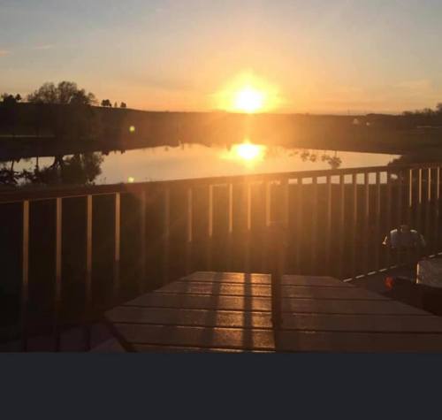 A serene sunset over a calm lake, with reflections on the water and silhouettes of trees in the background.