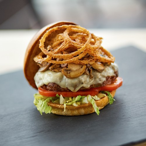 A delicious burger topped with melted cheese, sautéed mushrooms, crispy onion rings, lettuce, and tomato on a bun.