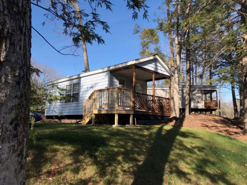 A white cabin with a wooden deck sits among trees on a grassy slope under a clear blue sky.