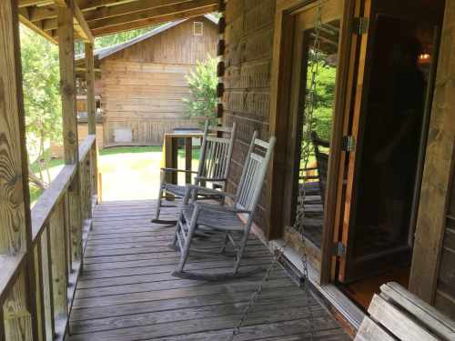 A wooden porch with two rocking chairs and a door leading inside, surrounded by greenery and rustic buildings.