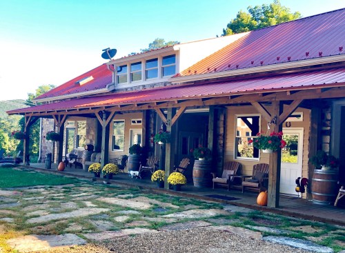 A charming rustic building with a red roof, wooden porch, and flower pots, surrounded by greenery and autumn decor.