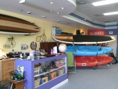 A colorful shop interior featuring kayaks, paddleboards, and bikes, with a reception area and various sporting goods.