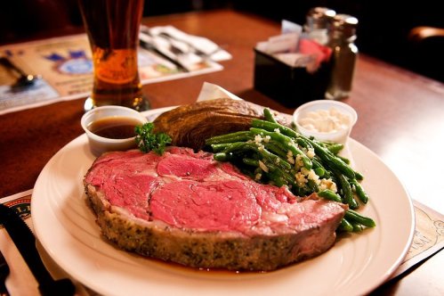 A plate of medium-rare prime rib with green beans, a baked potato, and dipping sauce, served in a restaurant setting.