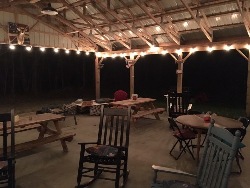 A rustic outdoor gathering space with wooden tables, rocking chairs, and string lights, set in a dark, wooded area.