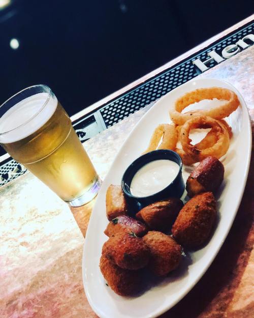 A plate of fried snacks including onion rings and potato bites, served with a dipping sauce, alongside a glass of beer.