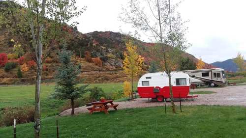 A red and white vintage trailer parked near a picnic table, surrounded by colorful autumn trees and mountains.