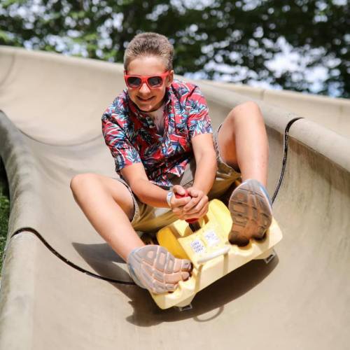 A young boy in a colorful shirt and sunglasses slides down a smooth slope on a yellow sled, smiling with excitement.