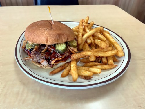 A plate with a pulled pork sandwich topped with pickles and a side of golden French fries.