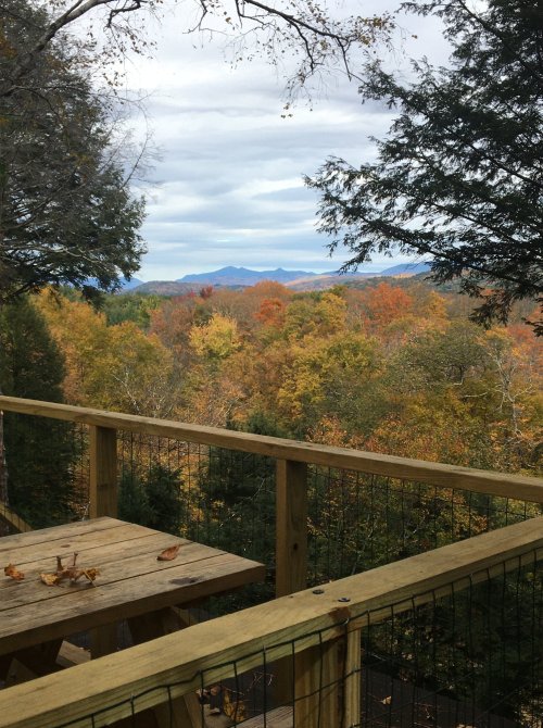 A wooden deck overlooks a vibrant autumn landscape with colorful trees and distant mountains under a cloudy sky.