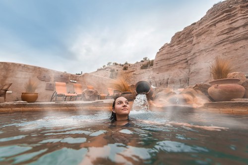 A woman relaxes in a hot spring, surrounded by rocky cliffs and steam rising from the water.