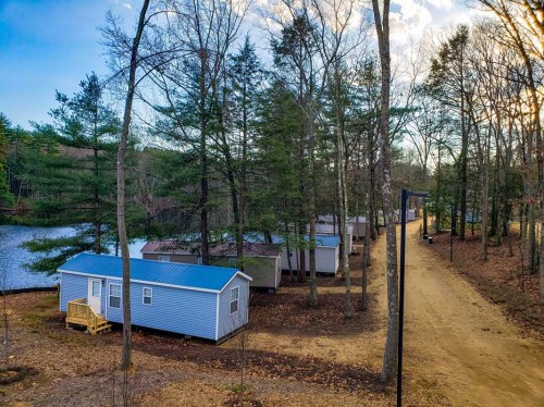 A row of blue cabins nestled among trees by a calm lake, with a dirt path leading through the serene landscape.
