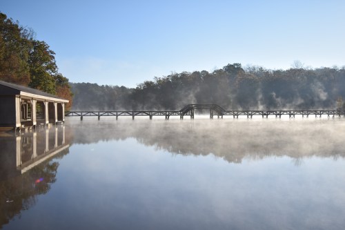 A serene lake scene with mist rising, a wooden dock, and trees in the background under a clear blue sky.