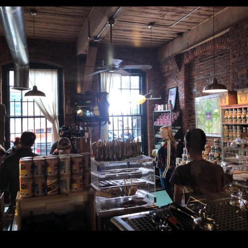 A cozy café interior with patrons, shelves of products, and large windows letting in natural light.