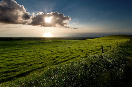 A vibrant landscape with green fields under a dramatic sky, featuring sunlight breaking through clouds at sunset.