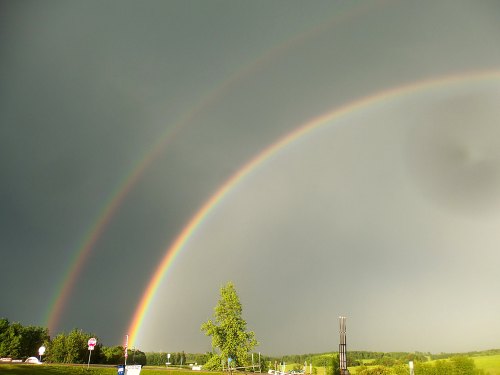 A vibrant double rainbow arcs across a dark, cloudy sky over a green landscape.