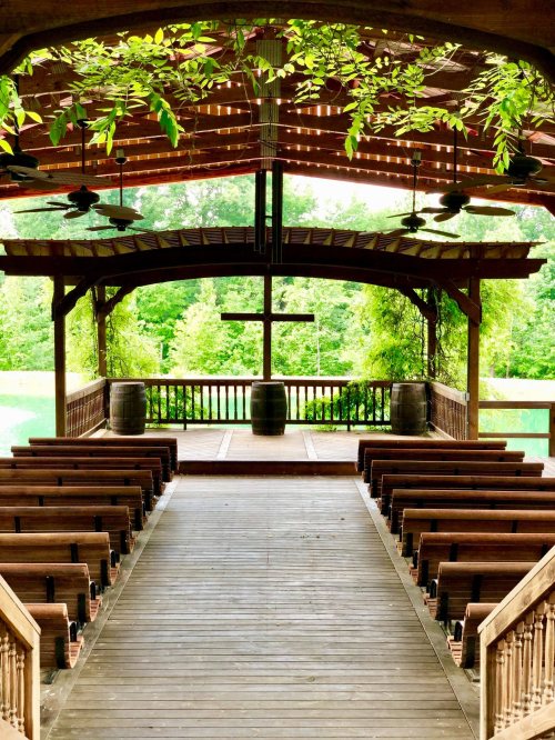 A wooden outdoor chapel with benches facing a cross, surrounded by greenery and natural light.