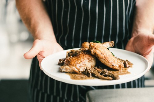 A chef in a striped apron holds a plate of roasted chicken with mushrooms and sauce, garnished with greens.