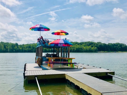 A colorful dock with umbrellas and a picnic table, surrounded by calm water and green trees under a blue sky.
