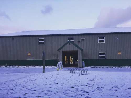 A snowy scene of a large, green and gray building with an entrance and bike rack in front.