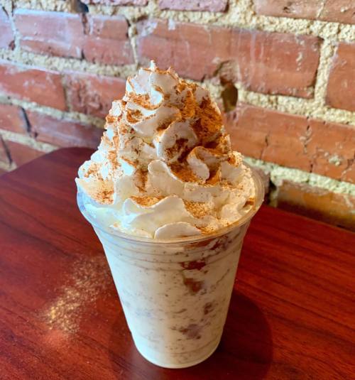 A creamy dessert in a cup topped with whipped cream and a sprinkle of cinnamon, set against a brick wall.