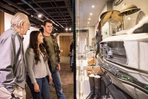 A group of three people observes exhibits in a museum, featuring various artifacts and a vintage vehicle.