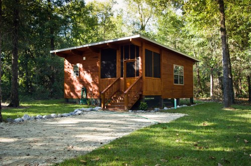 A wooden cabin surrounded by trees, featuring a porch and a gravel path leading to the entrance.