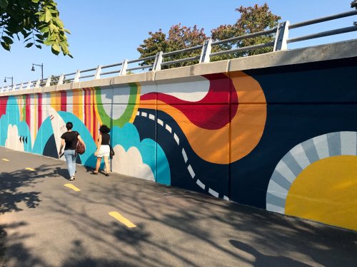 Two people walk along a path beside a colorful mural featuring abstract shapes and bright colors.