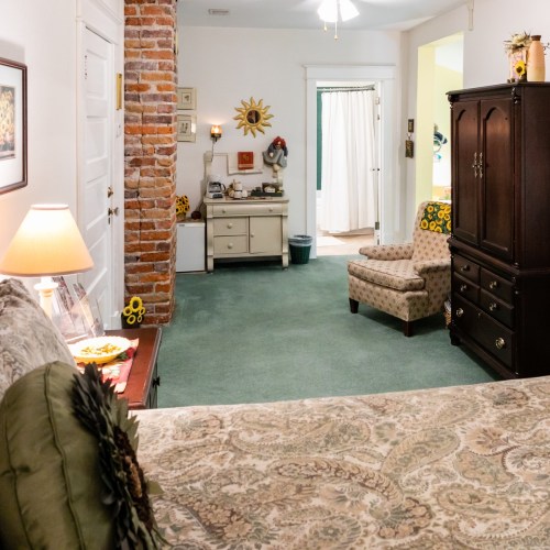 Cozy bedroom with a patterned bedspread, a brick wall, and a small sitting area featuring a chair and dresser.