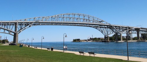 A large steel bridge spans over a calm body of water, with a grassy area and walking path in the foreground.