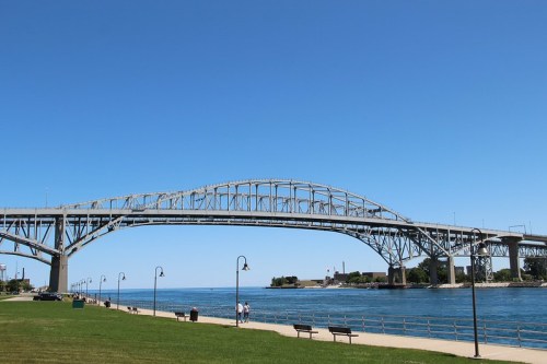 A large steel bridge spans over a calm river, with a clear blue sky and green grass in the foreground.