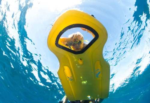 A person underwater looking through a yellow snorkel board, surrounded by clear blue water and sunlight.