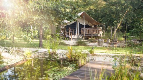 A cozy tent cabin surrounded by greenery and a tranquil pond, with a wooden deck and pathway.