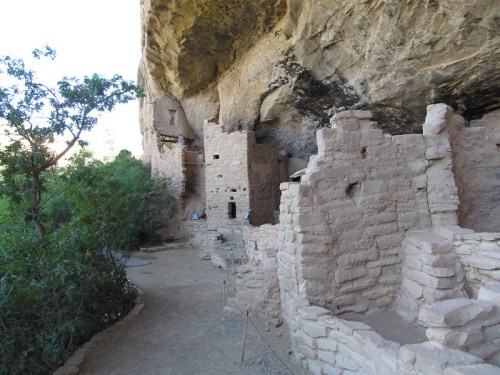 Ancient stone ruins nestled under a rocky overhang, surrounded by greenery and a dirt path.