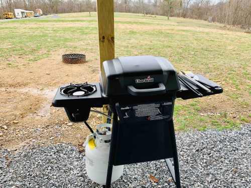 A black gas grill with a side burner on a gravel patio, set against a grassy field and trees in the background.