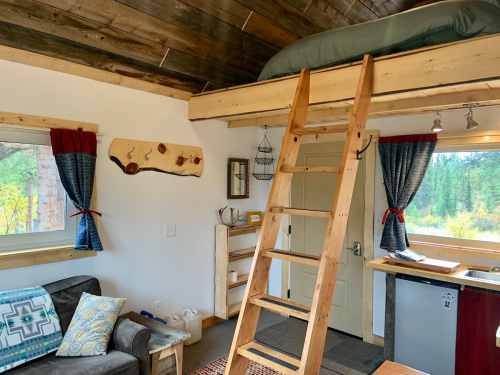 Cozy interior of a cabin with a loft bed, wooden ladder, and rustic decor, featuring a small kitchen and large windows.