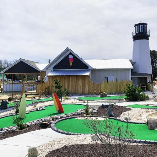 A mini-golf course with colorful surfboards in front of a building featuring a lighthouse and an ice cream cone sign.