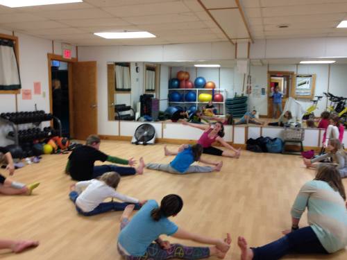 A group of children stretching in a fitness studio with exercise equipment and mirrors in the background.