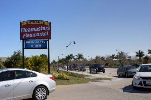 Sign for Fleamasters Fleamarket with cars parked and palm trees in the background under a clear blue sky.